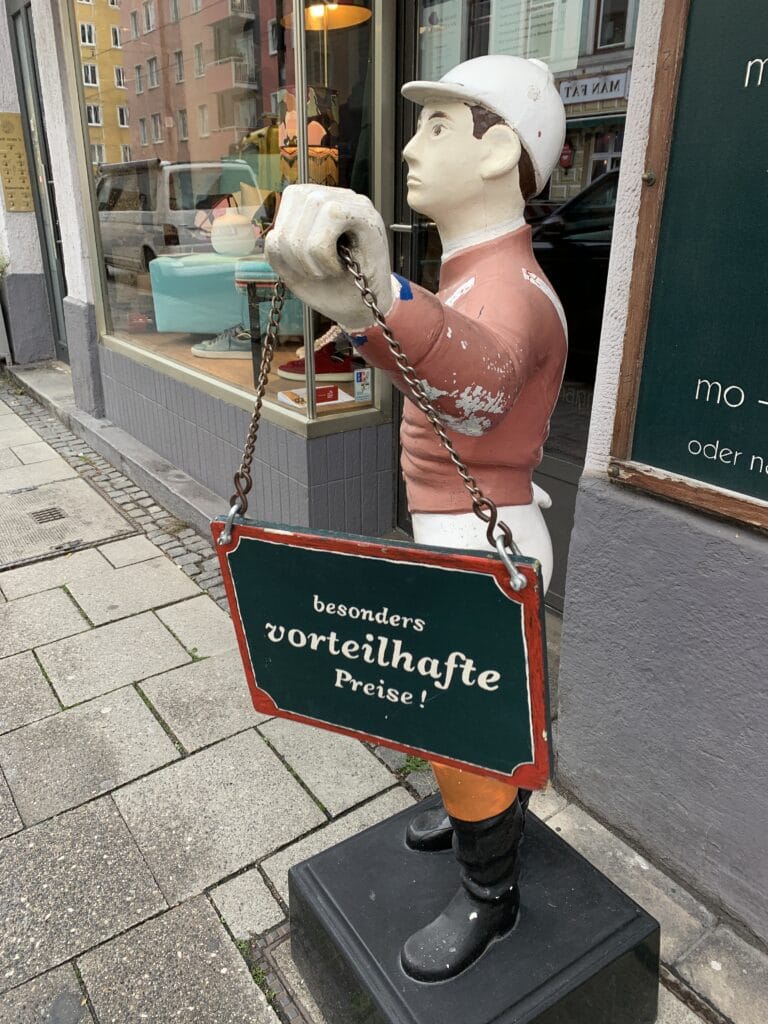 Historische Werbefigur mit Schild „besonders vorteilhafte Preise!“ vor dem Eingang von Second Gerdismann in der Münchner Maxvorstadt. Der Secondhand-Store bietet hochwertige Herrenmode, Designermarken und sorgfältig ausgewählte Vintage-Klassiker.