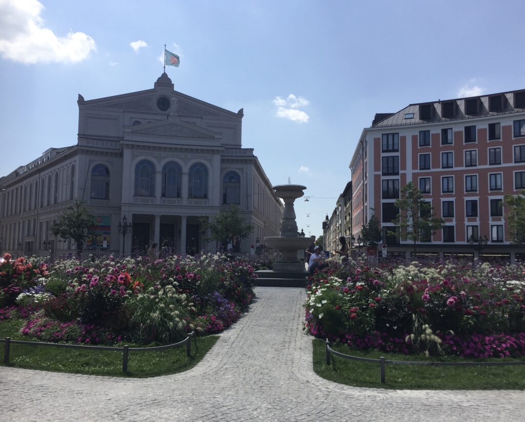 Blick auf den Gärtnerplatz in München mit Brunnen, Altbauten und Blumen im beliebten Glockenbachviertel.