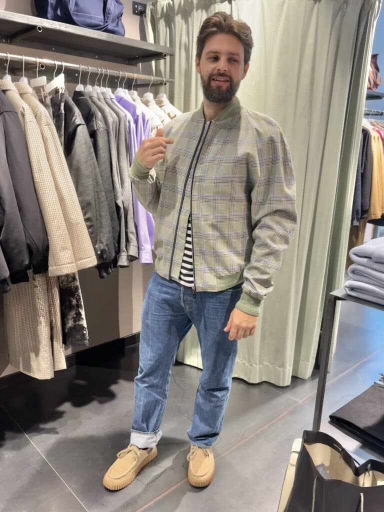 Kunde probiert im Store von J'ai mal à la tête in München eine karierte Bomberjacke an. Das Münchner Label ist bekannt für hochwertige Herrenmode mit modernen Schnitten und außergewöhnlichen Materialien.